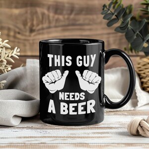 Könnte beinhalten: Schwarze Keramik-Tasse mit dem weißen Text "THIS GUY NEEDS A BEER". Das Design beinhaltet weiße Handgesten. Die Tasse hat einen schwarzen Henkel und steht vor einem Holzgrund.