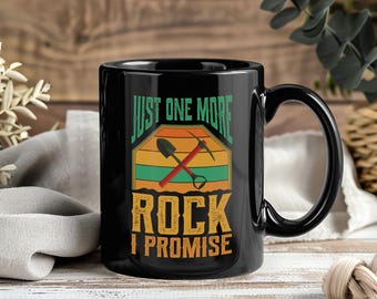 May include: Black ceramic mug with the text "JUST ONE MORE ROCK I PROMISE" in green and orange. The mug features a graphic of a shovel and pickaxe. The mug has a black handle and is on a wooden surface.
