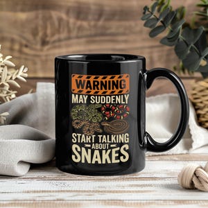 May include: A black ceramic mug featuring an orange and black hazard warning graphic at the top. The text reads WARNING MAY SUDDENLY START TALKING ABOUT SNAKES. Below the text are illustrations of four different snakes in green, red, and brown.