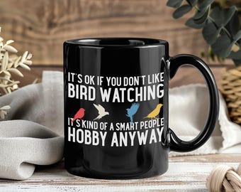 May include: Black ceramic mug with a curved handle. The mug features the text "IT'S OK IF YOU DON'T LIKE BIRD WATCHING" and "IT'S KIND OF A SMART PEOPLE HOBBY ANYWAY" in white. Colorful bird illustrations are also included.