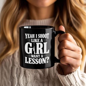 May include: A black ceramic mug featuring white text that reads YEAH I SHOOT LIKE A GIRL WANT A LESSON? The letter I in the word GIRL is replaced by a graphic of a rifle. The mug has a curved handle on the right side and a smooth finish.