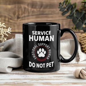 May include: Black ceramic mug with the text "SERVICE HUMAN" above "EMOTIONAL SUPPORT HUMAN" surrounding a paw print, and "DO NOT PET" below. The mug has a black handle and is on a wooden surface.