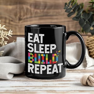 Könnte beinhalten: Schwarze Keramik-Tasse mit den Worten "EAT SLEEP BUILD REPEAT" in Weiß. Das Wort "BUILD" ist in bunten Buchstaben gehalten. Die Tasse hat einen großen Henkel und steht auf einer Holzoberfläche.