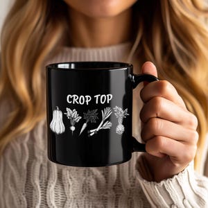 May include: Black ceramic mug with the words "CROP TOP" in white, along with illustrations of various vegetables. The mug is held by a person with long blonde hair.