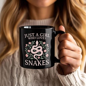 Puede incluir: Una taza de cerámica negra con un gráfico de una serpiente de color rosa claro enroscada, rodeada de flores rosas y hojas verdes. El texto impreso en rosa claro dice "JUST A GIRL WHO LOVES SNAKES", con la ilustración de la serpiente en el centro.