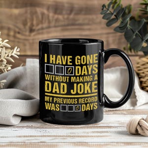 May include: Black ceramic mug with a large handle. The mug features the text "I HAVE GONE 0 DAYS WITHOUT MAKING A DAD JOKE" in yellow and white lettering. The mug is on a wooden surface.