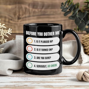May include: Black ceramic mug with a curved handle. The mug features the text "BEFORE YOU BOTHER ME" and a checklist: "1. IS IT PLUGGED IN?", "2. IS IT TURNED ON?", "3. ARE YOU SURE?", and "4. SERIOUSLY, GO CHECK".