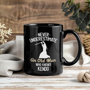 May include: Black ceramic mug with a white graphic of a kendo fighter and the text "NEVER UNDERESTIMATE An Old Man WHO KNOWS KENDO". The mug has a black handle and a glossy finish. The mug is on a wooden surface.