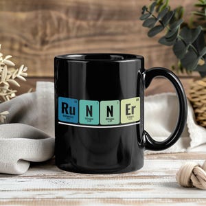 Puede incluir: Una taza de cerámica negra con la palabra "RUNNER" escrita con elementos de la tabla periódica. Las letras están en cajas azules, verdes y amarillas. La taza tiene un asa negra y está sobre un fondo de madera.