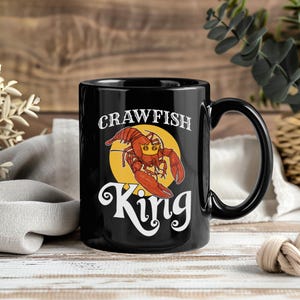 May include: Black ceramic mug with a red and yellow crawfish design. The mug features the words "CRAWFISH" above the crawfish and "King" below it, in a white, stylized font. The mug has a black handle.