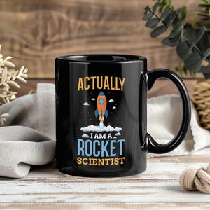 May include: Black ceramic mug with the text "ACTUALLY I AM A ROCKET SCIENTIST" in orange and blue. A cartoon rocket ship graphic is in the center. The mug has a black handle and is on a wooden surface.