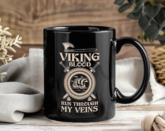 May include: A black ceramic mug with a curved handle. The mug features the text "VIKING BLOOD RUN THROUGH MY VEINS" in a beige font, along with a Viking ship and axe design. The mug is on a wooden surface.