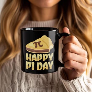 Puede incluir: Taza de cerámica negra con un gráfico de una porción de tarta, el símbolo pi y las palabras "HAPPY PI DAY" en amarillo. La taza tiene un asa negra. La taza está sujeta por una persona.