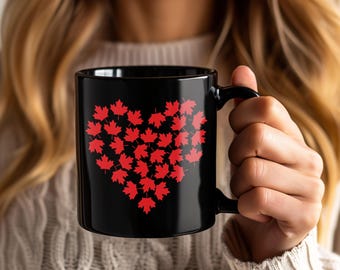 May include: Black ceramic mug with a heart design composed of red maple leaves. The mug has a black handle and is held by a person wearing a cream-coloured jumper. The design is a symbol of Canada.