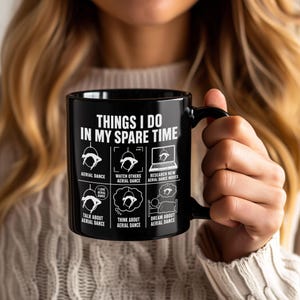 May include: A black ceramic mug featuring white text and graphics. The text reads: THINGS I DO IN MY SPARE TIME, followed by six panels illustrating aerial dance activities: aerial dance, watch others, research moves, talk, think, and dream.
