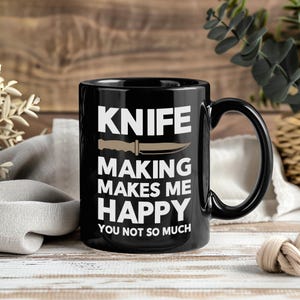 May include: Black ceramic mug with the text "KNIFE MAKING MAKES ME HAPPY YOU NOT SO MUCH" in white. A tan knife graphic is centered above the text. The mug has a black handle and is on a wooden surface.