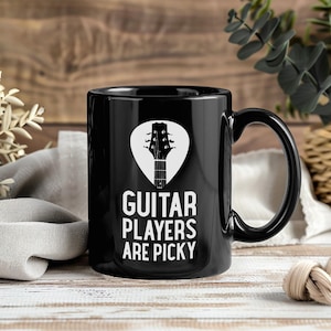 Può includere: Tazza da caffè in ceramica nera con grafica bianca di una paletta di chitarra all'interno di un plettro. La tazza presenta la scritta "GUITAR PLAYERS ARE PICKY" in lettere bianche. La tazza ha un manico nero.
