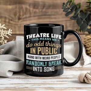 Op de afbeelding: Zwarte keramische mok met de tekst "THEATRE LIFE" en "do odd things IN PUBLIC". De mok zegt ook "HANG WITH WEIRD PEOPLE" en "RANDOMLY BREAK INTO SONG". De mok heeft een zwart handvat.