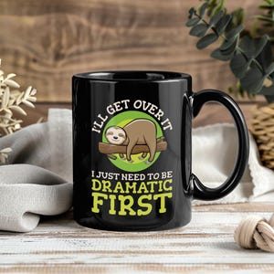 Puede incluir: Taza de cerámica negra con una ilustración de un perezoso y el texto "I'll get over it, I just need to be dramatic first." El perezoso es marrón y descansa sobre una rama marrón, sobre un círculo verde.