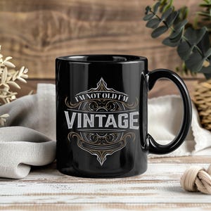 Może przedstawiać: Czarny ceramiczny kubek z dużym uchwytem. Kubek zawiera napis "I'M NOT OLD I'M VINTAGE" w ozdobnej, stylizowanej czcionce. Projekt obejmuje bogate zdobienia i klasyczną estetykę, odpowiednią do napojów.
