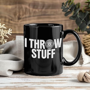 Puede incluir: Taza de cerámica negra con el texto blanco "I THROW STUFF" y un gráfico de disco. La taza tiene un asa curva y está sobre un fondo de madera.