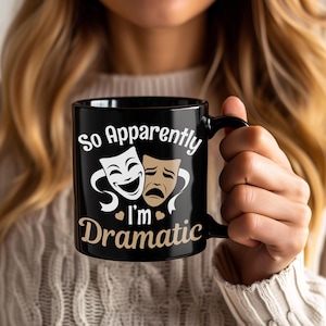 Könnte beinhalten: Schwarze Keramik-Tasse mit dem Text "So Apparently I'm Dramatic" in weißer und goldener Schrift. Die Tasse zeigt ein Design mit einem lächelnden und einem verärgerten Theatermasken-Motiv. Die Tasse wird von einer Person gehalten.