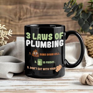 May include: Black ceramic mug with the text "3 Laws of Plumbing" and humorous illustrations. The mug features the phrases "Runs Down Hill," "Is Friday," and "Don't Eat With Your." The mug has a black handle.