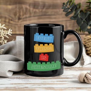 May include: Black ceramic mug featuring the phrase "EVERYTHING IS AWESOME" in a colourful block design. The text is arranged in a stacked format, with each word on a separate block. The mug has a large handle and is set against a wooden background.