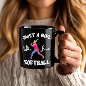 Puede incluir: Taza de cerámica negra con el texto blanco "JUST A GIRL Who Loves SOFTBALL". La taza presenta una ilustración de acuarela colorida de una jugadora de softbol. La taza es sostenida por una persona.