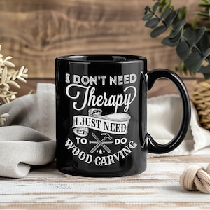 May include: Black ceramic mug with the text "I DON'T NEED Therapy I JUST NEED TO DO WOOD CARVING" in white. The mug features a black handle and a humorous message for woodworkers. The mug is on a wooden surface.