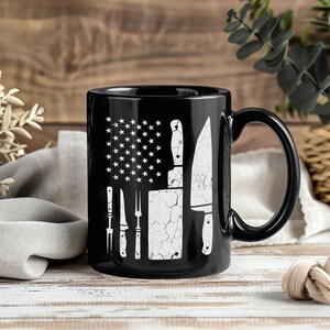 May include: Black ceramic mug featuring a white graphic of the American flag with kitchen knives and utensils replacing the stripes and stars. The mug has a black handle and is suitable for hot or cold beverages.