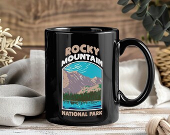 May include: Black ceramic mug featuring a scenic illustration of the Rocky Mountains National Park. The design includes a mountain range, lake, and trees under a blue sky. The words "Rocky Mountain National Park" are printed on the mug.