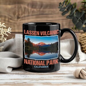 May include: Black ceramic mug featuring a scenic image of Lassen Volcanic National Park in California. The mug has a large handle and the park's name is printed in orange. The image shows a lake and mountains.