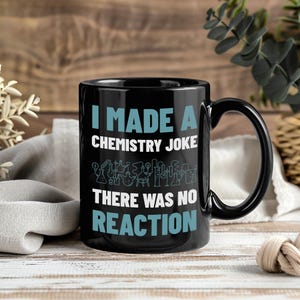 以下が含まれることがあります： 「I MADE A CHEMISTRY JOKE THERE WAS NO REACTION」の文字が白とターコイズブルーでプリントされた黒いセラミックマグ。マグには化学関連のイラストが描かれています。マグは黒いハンドル付きで、木製の背景に置かれています。
