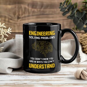 Könnte beinhalten: Schwarze Keramik-Tasse mit dem Text "ENGINEERING SOLVING PROBLEMS" in Gelb. Darunter befindet sich eine gelbe Grafik eines Gehirns aus Leiterbahnen. Auf dem Boden der Tasse steht der Text "YOU DIDN'T KNOW YOU HAD IN WAYS YOU CAN'T UNDERSTAND."