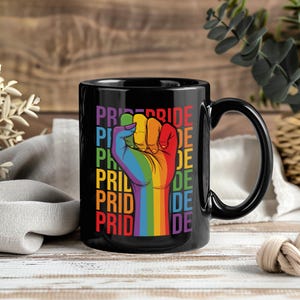 Op de afbeelding: Zwarte keramische mok met een regenboogvuistafbeelding en het woord "PRIDE" in regenboogkleuren. De mok heeft een zwart handvat en staat tegen een houten achtergrond.