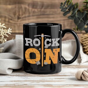 May include: Black ceramic mug with the words "ROCK ON" in white and orange, with a pickaxe graphic. The letter "O" is designed to look like cracked rock. A great gift for a geologist or rock enthusiast.