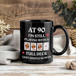 May include: Black ceramic mug with a curved handle. The mug features the text "AT 90 I'M STILL PLAYING WITH A FULL DECK I JUST SHUFFLE SLOWER" in white, along with playing card suit symbols and card images.
