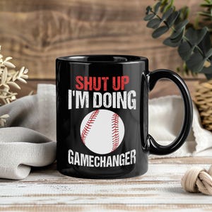 Könnte beinhalten: Schwarze Keramik-Tasse mit dem Text "SHUT UP I'M DOING GAMECHANGER" in Weiß und Rot. Eine Baseball-Grafik befindet sich in der Mitte. Die Tasse hat einen schwarzen Griff und steht auf einer Holzoberfläche.