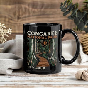 May include: Black ceramic mug featuring a scenic illustration of Congaree National Park in South Carolina. The design includes a wooden walkway through a forest, with the park's name and state printed on the mug.
