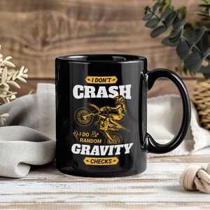 以下が含まれることがあります： 大きなハンドルが付いた黒いセラミックマグカップ。「I DON'T CRASH, I DO RANDOM GRAVITY CHECKS」の文字が白と黄色で表示され、オフロードバイクとライダーのグラフィックが描かれています。