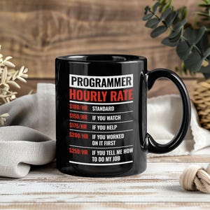 Könnte beinhalten: Eine schwarze Keramiktasse mit großem Henkel. Die Tasse zeigt den Text "PROGRAMMER HOURLY RATE" in Weiß und Rot sowie verschiedene Stundensätze, die sich nach dem Grad der geleisteten Hilfe richten. Die Tasse steht auf einer Holzoberfläche.