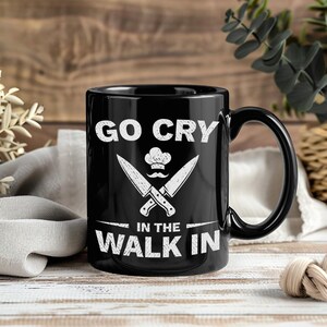 Könnte beinhalten: Schwarze Keramik-Tasse mit dem weißen Text "GO CRY IN THE WALK IN". Das Design umfasst gekreuzte Messer, eine Kochmütze und einen Schnurrbart. Die Tasse hat einen schwarzen Griff und steht auf einer Holzoberfläche.