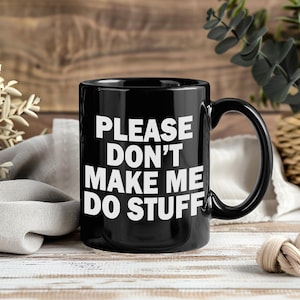 May include: Black ceramic mug with the text "PLEASE DON'T MAKE ME DO STUFF" in white block letters. The mug has a large handle and is set against a blurred background with greenery and a wooden surface.