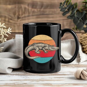 May include: Black ceramic mug with a retro design. The mug features an illustration of an alligator set against a sunset with orange, yellow, red, and blue stripes. The mug has a large handle.