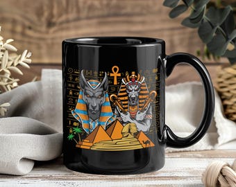 May include: Black ceramic mug featuring an Egyptian theme. The design includes illustrations of Anubis and a pharaoh, pyramids, and hieroglyphs. The mug has a black handle and a glossy finish, perfect for coffee or tea.