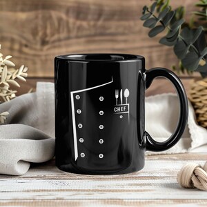 Könnte beinhalten: Schwarze Keramik-Tasse mit weißem Kochjacken-Design, einschließlich Knöpfen und dem Wort "CHEF" mit Besteck. Die Tasse hat einen gebogenen Griff und ist für heiße oder kalte Getränke geeignet.