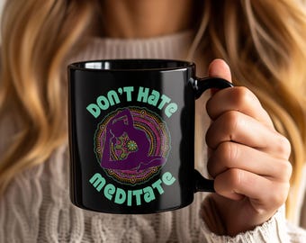 May include: Black ceramic mug with the words "DON'T HATE MEDITATE" in teal lettering. The mug features a purple yoga pose illustration within a colorful mandala design. The mug has a black handle.