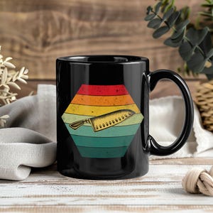 Könnte beinhalten: Schwarze Kaffeetasse mit einem Retro-Design eines Kochmessers in einem farbenfrohen Sechseck. Das Sechseck hat ein Streifenmuster mit roten, orangen, gelben, grünen und blauen Farbtönen. Die Tasse hat einen großen Henkel.