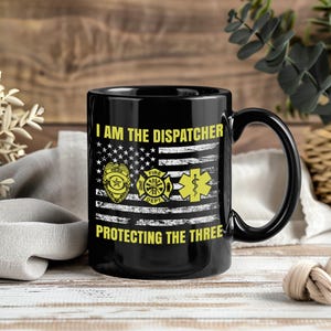 Puede incluir: Taza de café negra con el texto "I AM THE DISPATCHER PROTECTING THE THREE" y un diseño de bandera estadounidense. La bandera incorpora símbolos de policía, bomberos y médicos en amarillo. La taza tiene un asa negra.
